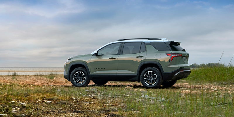 Side profile of a light green Chevrolet Equinox SUV parked on rocky grassland near a shoreline under a cloudy sky