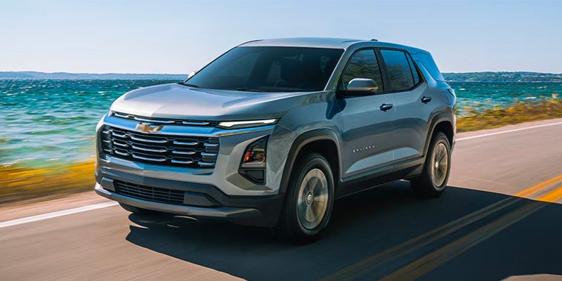 Silver Chevrolet Equinox SUV driving on a coastal road beside blue water on a sunny day, shown from the front three-quarter angle