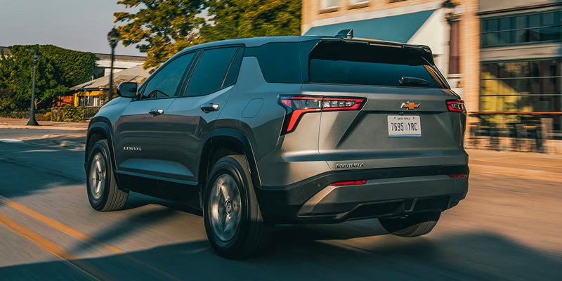 Rear three-quarter view of a silver Chevrolet Equinox SUV driving through an urban street, with buildings and trees blurred in the background