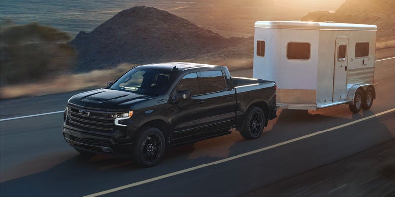 Black Chevrolet Silverado pickup truck towing a white enclosed trailer along a highway with mountains in the background at sunset