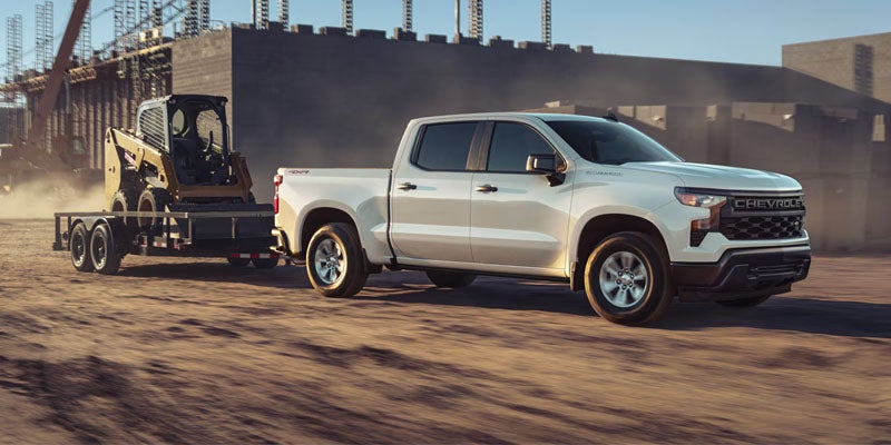 White Chevrolet Silverado pickup truck towing a trailer with a skid steer loader through a dusty construction site