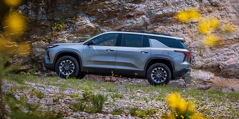 Side view of a gray SUV parked on a rocky, grassy terrain in front of a stone cliff