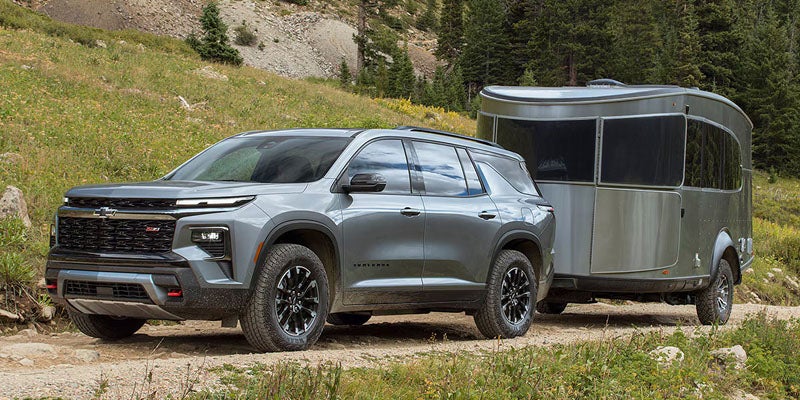 Gray SUV towing a silver travel trailer on a dirt road through a mountainous landscape