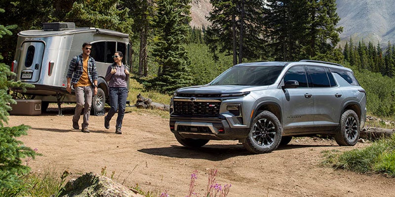 Gray SUV parked at a forest campsite beside a silver travel trailer on a dirt clearing