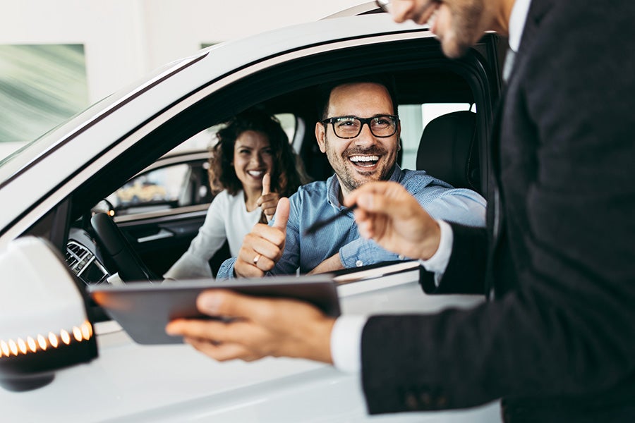 A customer seated inside a vehicle gives a thumbs-up while a salesperson holding a tablet stands beside the open window