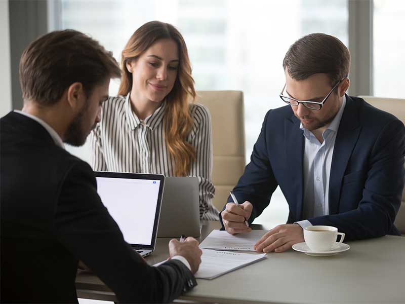 Three people seated around a meeting table reviewing and signing documents