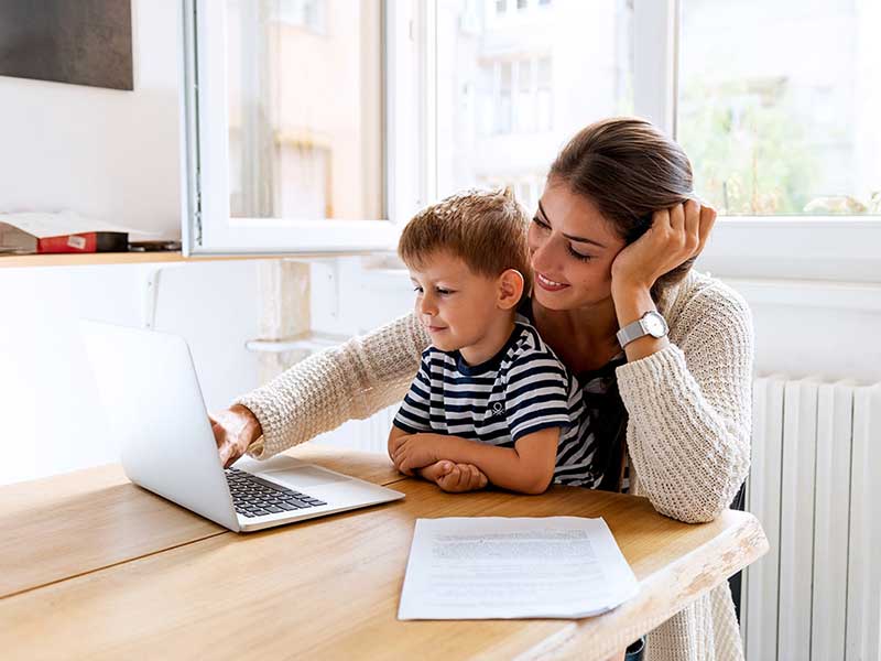 Person and child sitting together at a table, looking at a laptop screen in a bright home setting with documents on the table