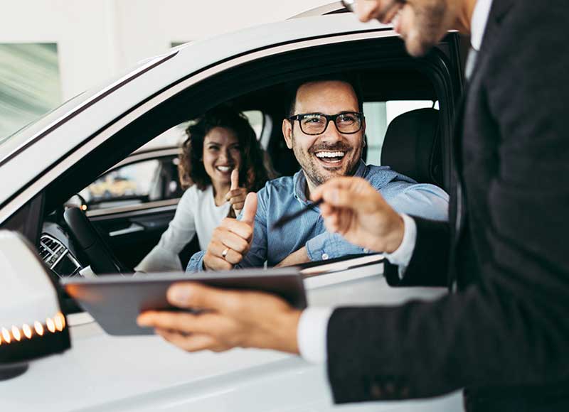 Two people seated inside a white car while a salesperson standing outside holds a tablet