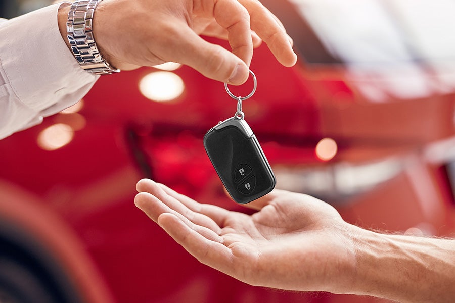 A close-up of a car key fob being handed from one person to another, with a red vehicle blurred in the background
