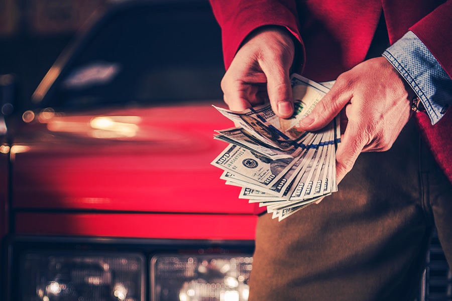 Close-up of a person holding and counting several U.S. dollar bills while standing in front of a red vehicle